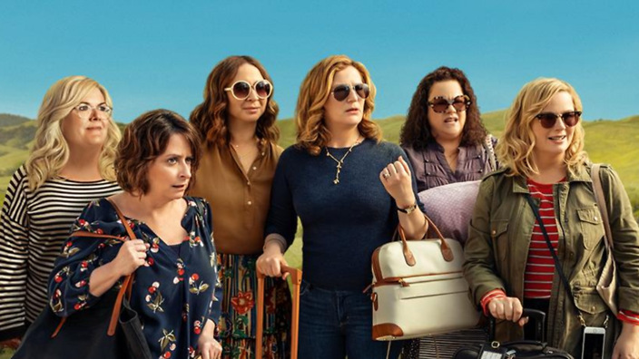 Cast from Wine Country stand on a green hill with their luggage