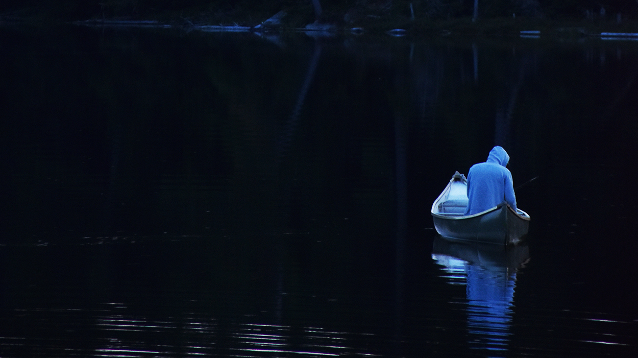 Hooded figure sits paddling in boat on the dark water with a mirror-like reflection in a cool blue on a lake at dusk or dawn