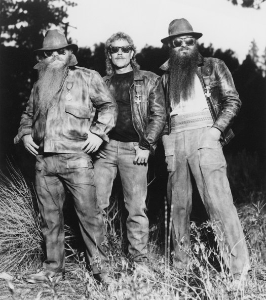 ZZ Top standing in the woods in tallish grass. They are wearing rustic clothes, vintage denim and leather jackets.