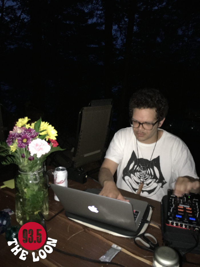 Dougie Boom DJing seated at a table in a dark forest. On the table are flowers and laptop that he is looking at.