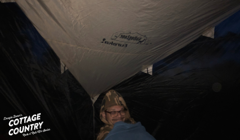 Man in camo hoody in sleeping bag, smiling and seated in the corner of a tent