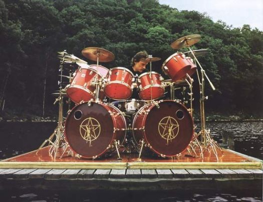 Neil Peart drumming outdoors on a dock on lake with trees in the background. The 2 kick drums have the Rush Star logo on it.