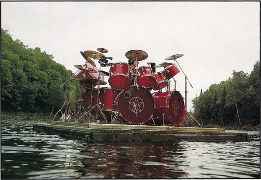 Neil Peart playing a full drum kit on a raft on a lake with surrounding trees