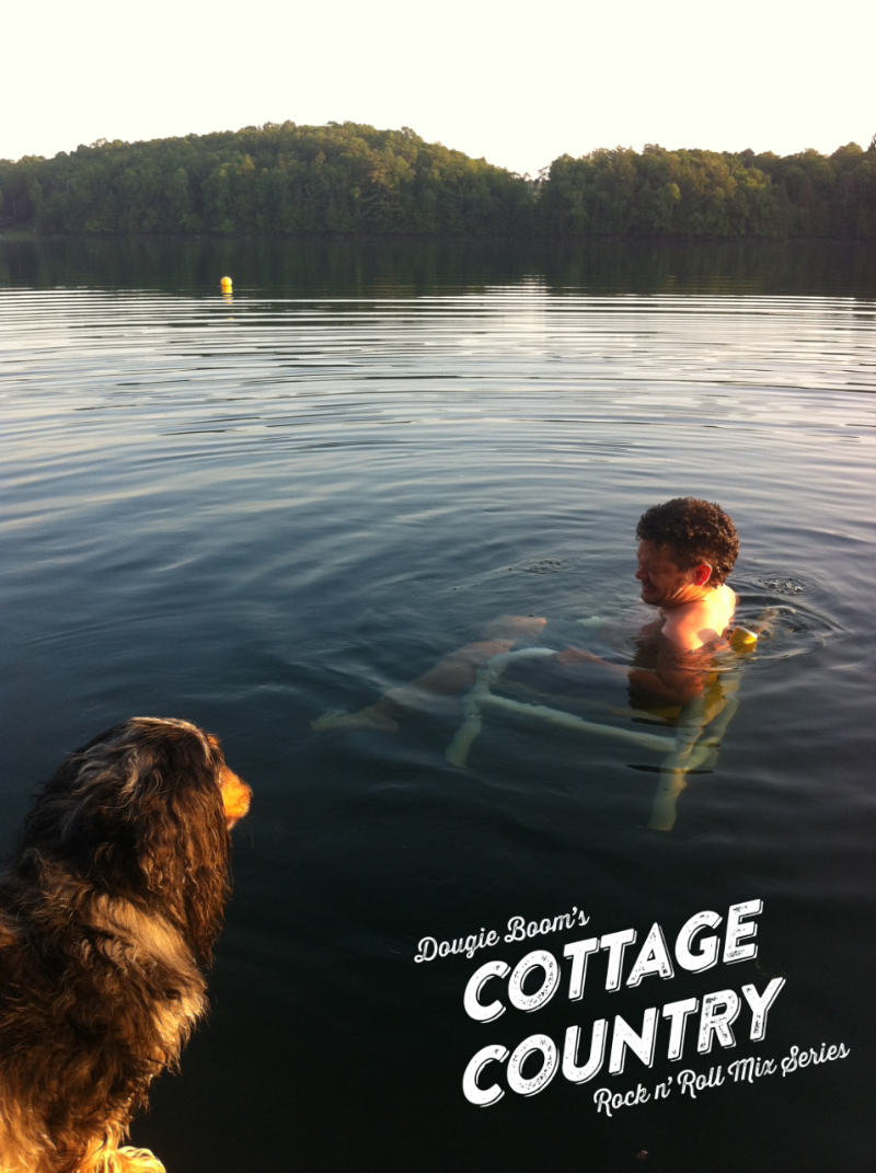 Someone sitting on a chair submerged in water on a lake with trees. A dog in the foreground stares at the man.