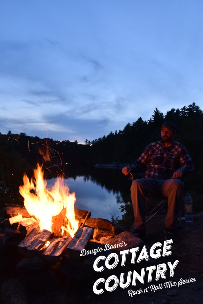 Man wearing flannel sits outdoors near a campfire. The man and forest is in the dark but the sky is a blue dusk.