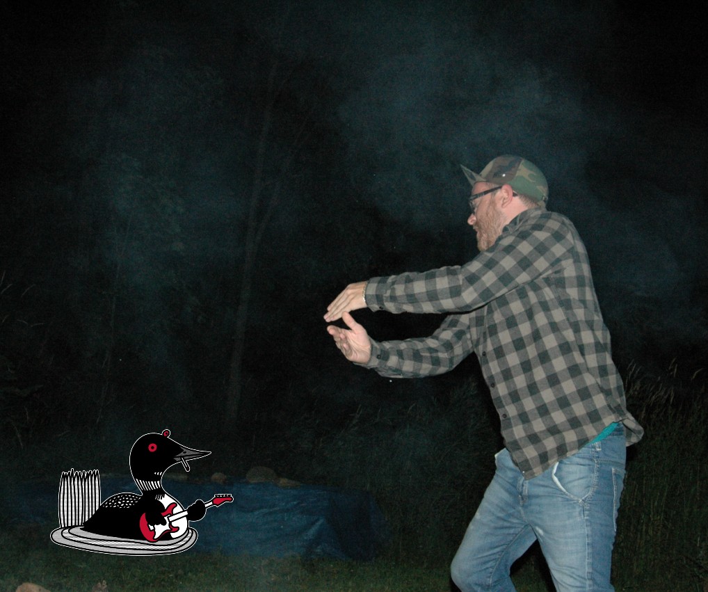 Man with flannel shirt and camo hat holds hands up to perform a charade in the dark night.